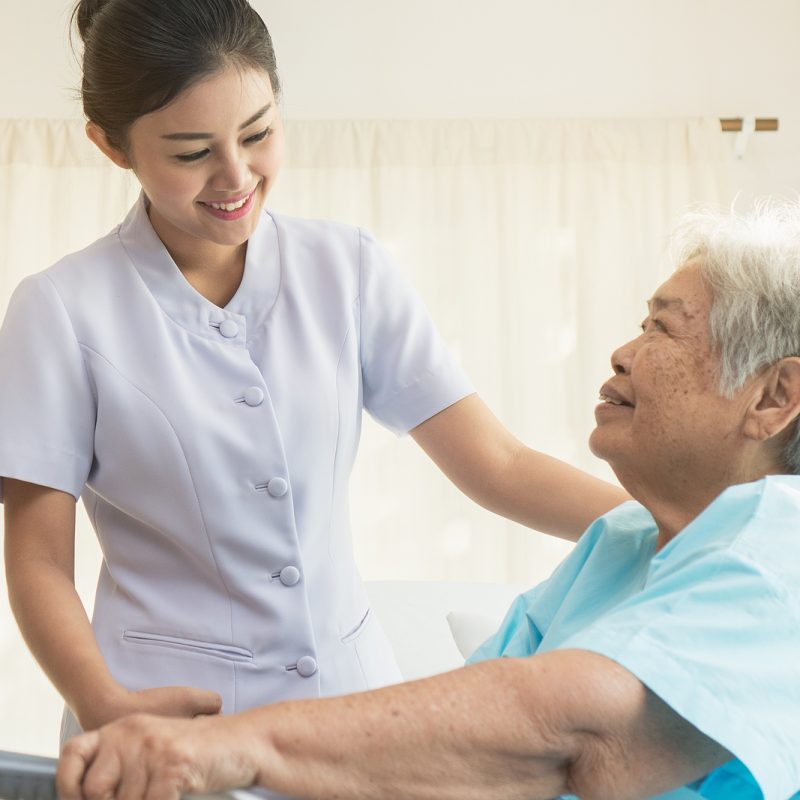 Cheerful asian nurse visiting elderly patient to check up after surgery in hospital for giving physical therapy and encouragement.